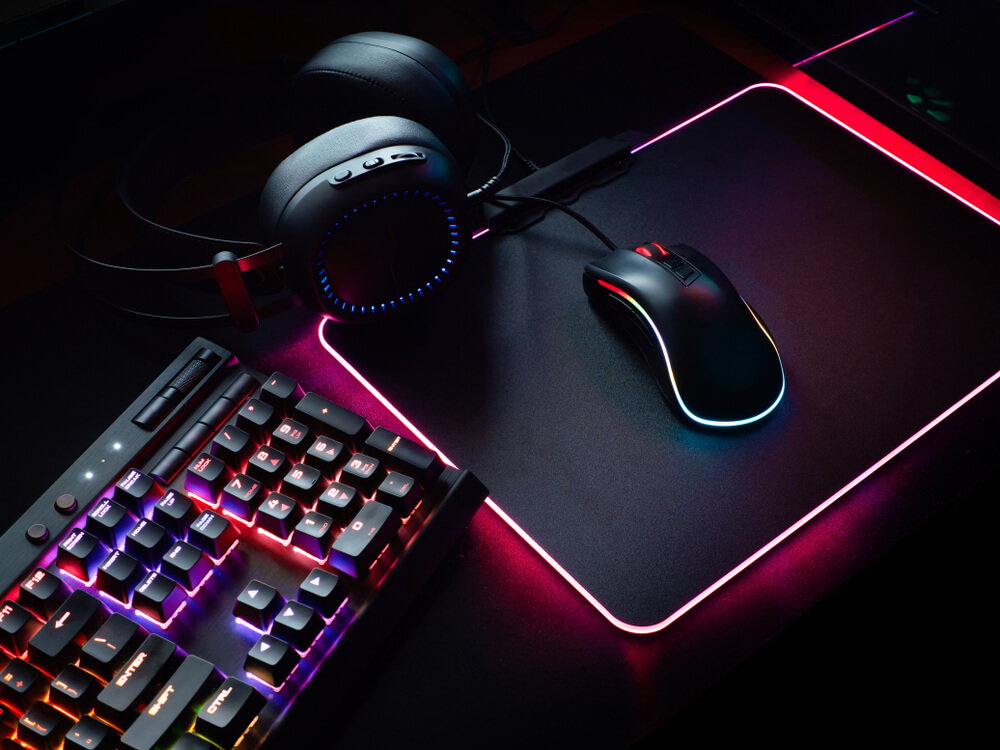 Foto que ilustra matéria sobre sala gamer mostra acessórios como teclado, fone, mouse e mousepad. Todos têm a cor preta e com iluminações internas em rosa, vermelho, lilás ou azul.