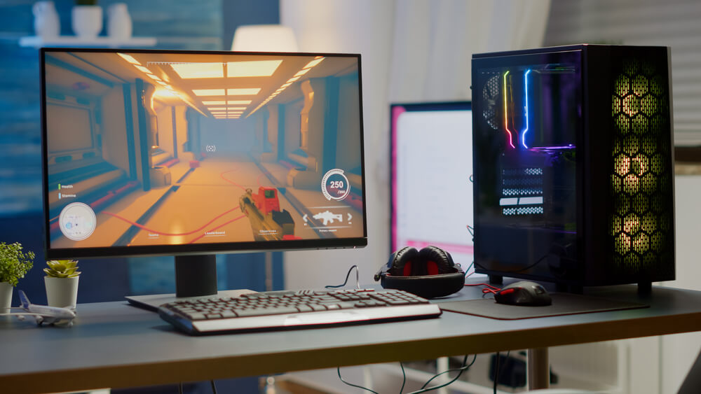Foto que ilustra matéria sobre sala gamer mostra uma mesa com computador, monitor, mouse, teclado e fones de ouvidos. Na tela, um jogo aparece.