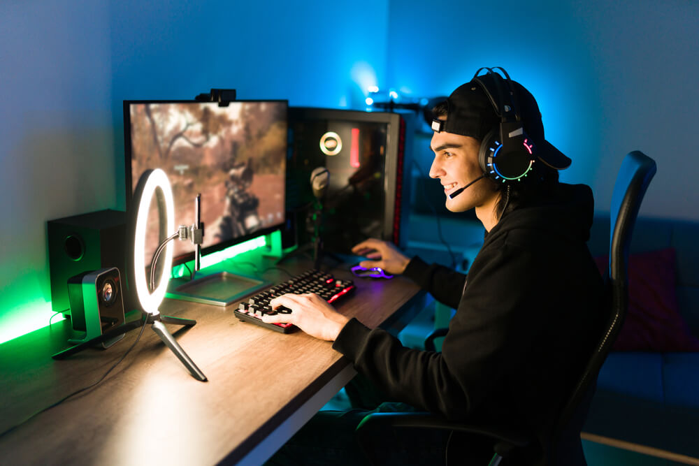 Foto que ilustra matéria sobre sala gamer mostra um jovem de perfil, sentado de frente para uma tela de computador jogando um game com uma das mãos em um teclado e outra no mouse. Na traseira da mesa, uma luz verde decora o ambiente.