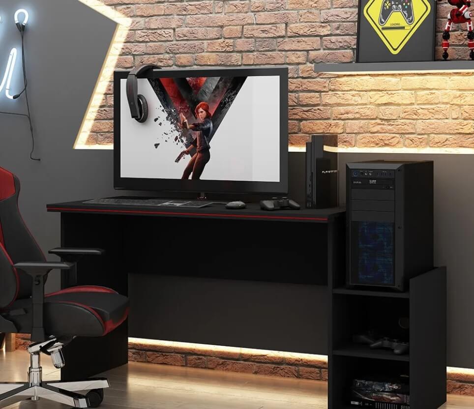 Foto que ilustra matéria sobre sala gamer mostra um mesa de computador toda preta, com o gabinete em uma estante anexa ao lado e um monitor acima com um game na tela.