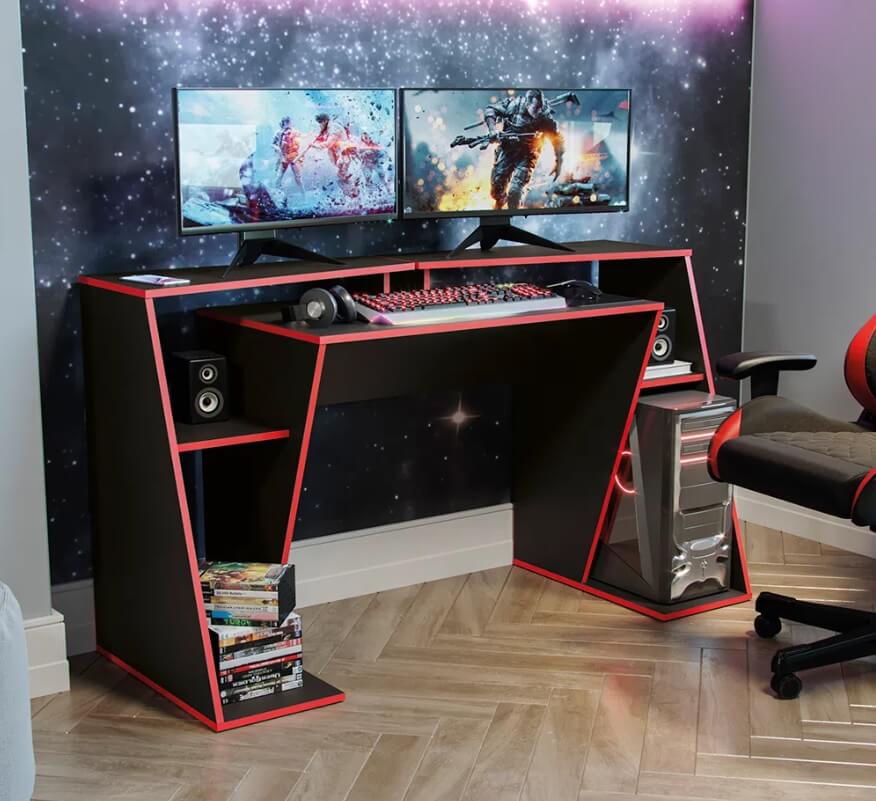 Foto que ilustra matéria sobre sala gamer mostra uma mesa preta, com detalhes em vermelho. Um gabinete de computador se encontra na parte lateral mais abaixo. Na parte de cima, há dois monitores com imagens de jogos. E em um nível mais abaixo, um espaço com fone, teclado e mouse. 