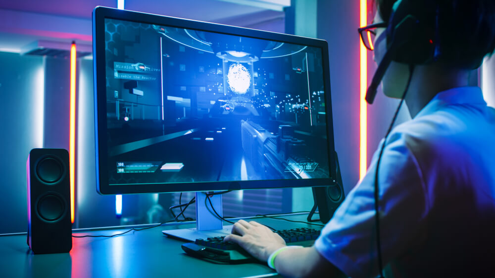 Foto que ilustra matéria sobre sala gamer mostra um jovem de óculos, de perfil, de frente para um grande monitor jogando um game com as mãos no teclado