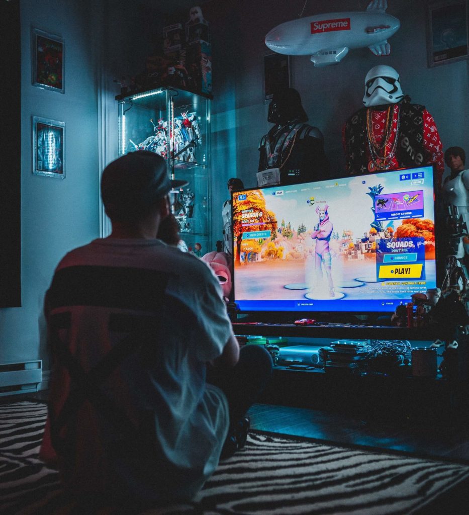 Foto que ilustra matéria sobre sala gamer mostra um homem de costas, sentado no chão em um ambiente escuro. Ele usa um boné. A sua frente uma grande TV com um jogo de videogame. O quarto é decorado com temas de Star Wars