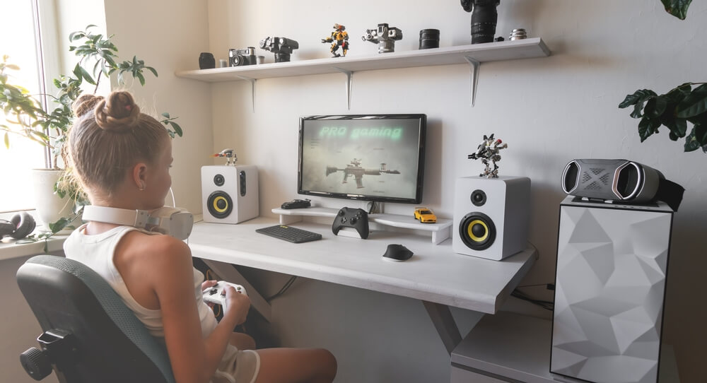 Foto que ilustra matéria sobre sala gamer mostra uma jovem sentada de frente para uma mesa encostada na parede com uma tela, caixas de som. O ambiente é todo branco e ela segura um controle enquanto olha para o jogo na tela.