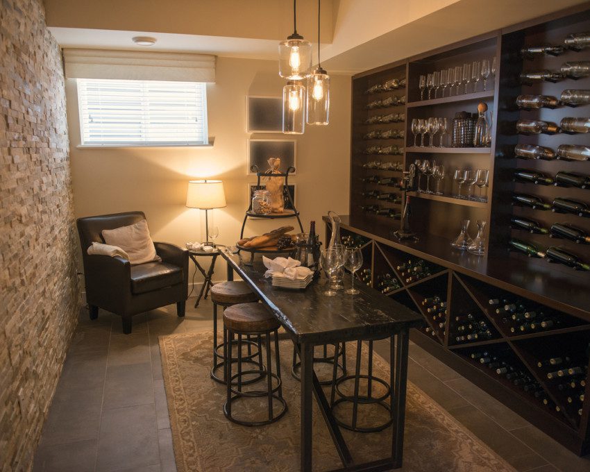 Adega com parede de vinhos à direita e poltronas à esquerda. No centro do ambiente, mesa com vinhos e taças