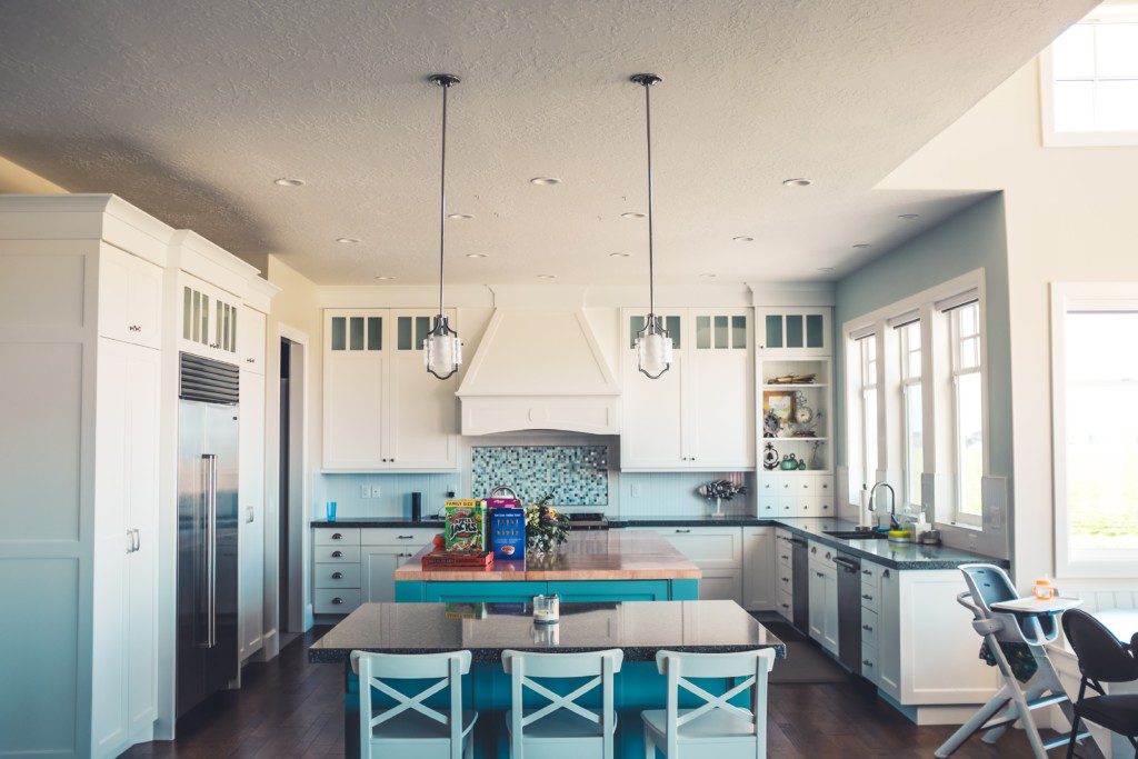 A foto mostra um exemplo de cozinha retrô em tons claros. A cozinha está completa com geladeira, armários em toda ela, mesa de centro com 3 cadeiras, uma bancada, pia e luminárias.