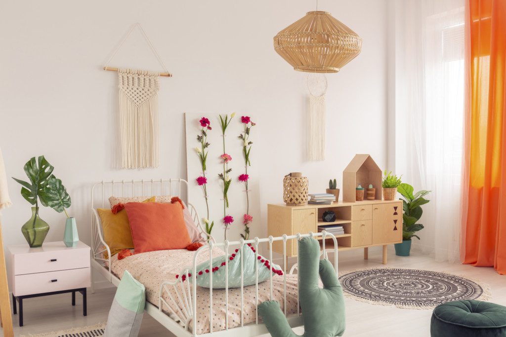 A imagem mostra um quarto com cama de solteiro, mesa de cabeceira e aparador. Há também elementos decorativos como plantas, tapete, puff, entre outros.