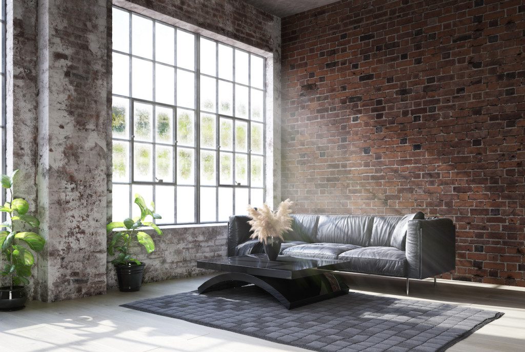  Na imagem há uma sala de estar com sofá, mesa de centro, tapete e diferentes plantas. As paredes do ambiente possuem tijolos aparentes, além de uma ampla janela.