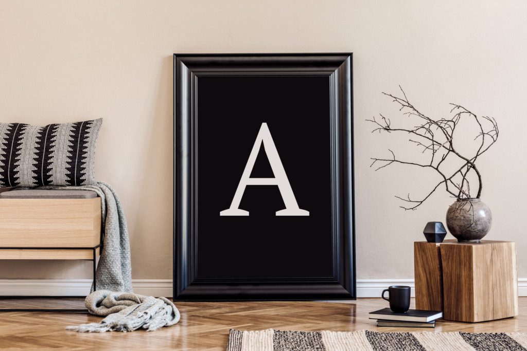 A imagem mostra um quadro com a letra A, um banco de madeira, um tapete e uma planta decorativa.