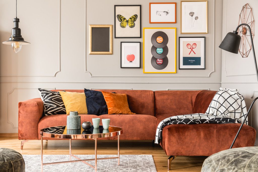 A imagem mostra uma sala de estar com sofá em L, mesa de centro, luminária de chão, lustre de teto e muitos quadros divertidos na parede.