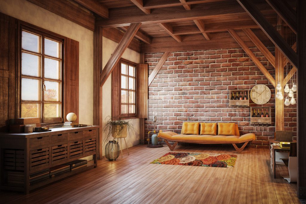 A imagem mostra um ambiente com um aparador, um sofá, tapete, plantas, parede de tijolo aparente e estantes com vinhos. 