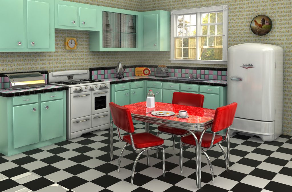 A imagem mostra uma cozinha com um armário em L. Nele há um fogão e uma pia embutidos. Tem também na imagem uma geladeira, uma mesa vermelha com 3 cadeiras ao redor e um piso de xadrez.