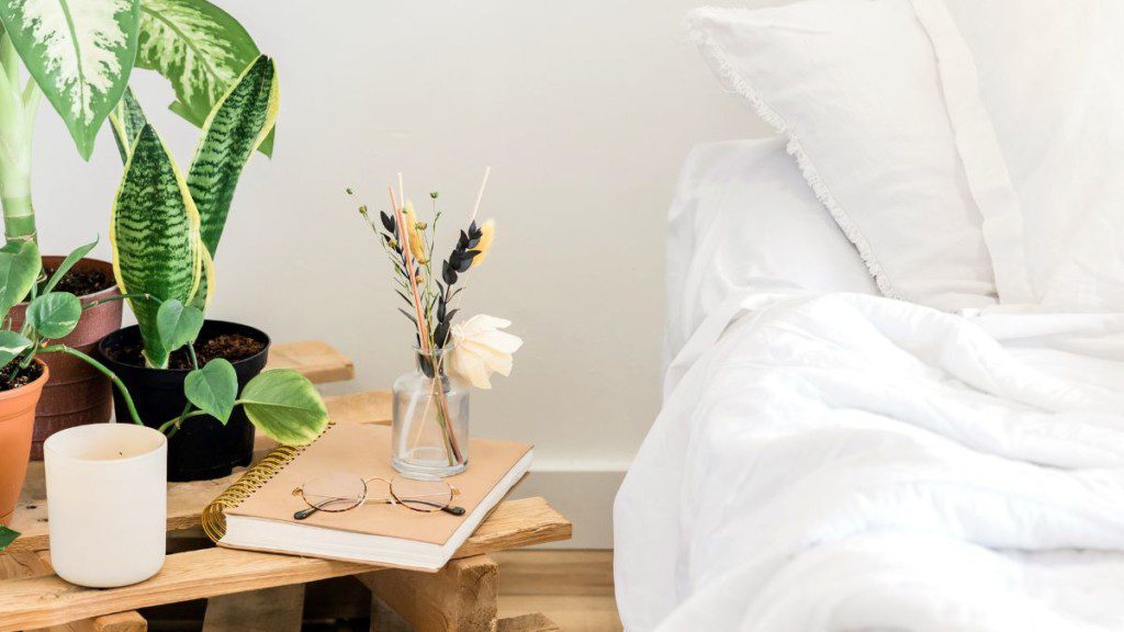 beira da cama com lençois brancos, ao lado de uma mesa de cabeceira de madeira com plantas em cima