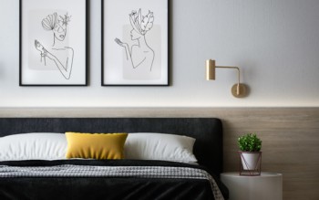 quarto com decoração minimalista. Cama com coberta preta e almofadas amarelas, quadros acima da cama com desenhos e pequena mesa com planta