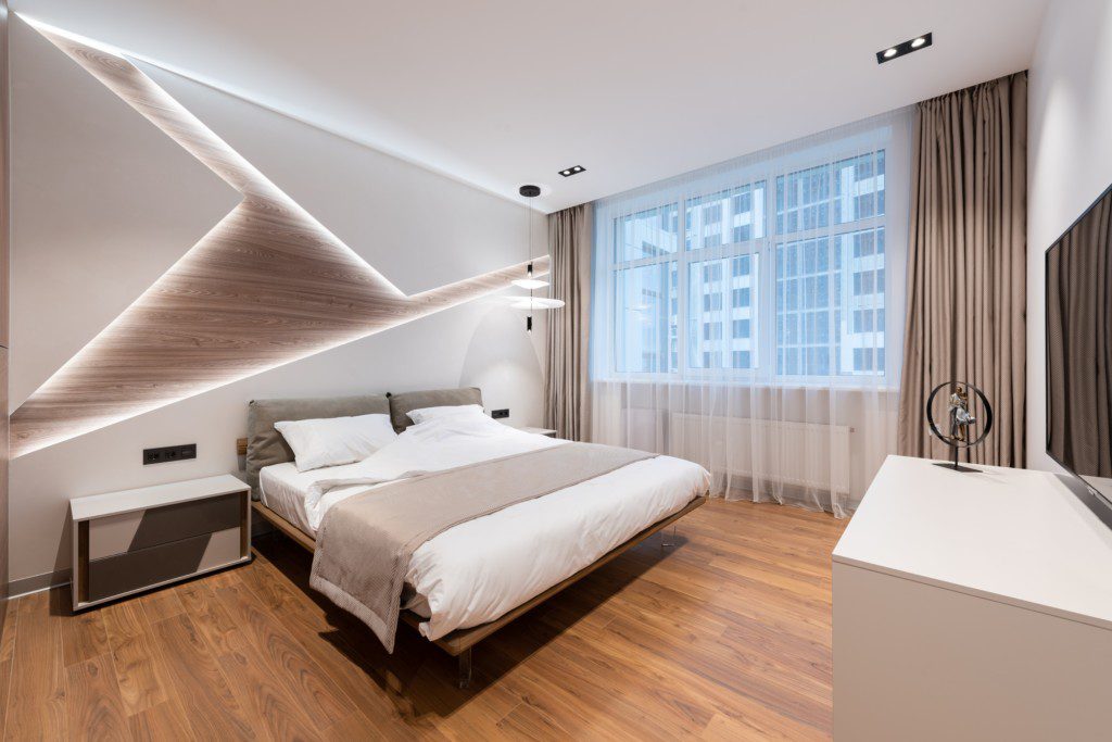 Foto de um quarto moderno com uma cama de casal baixa, uma mesa de cabeceira, cortina e cômoda com TV em cima. Há também um painel de formato abstrato feito em madeira e com iluminação ao redor.