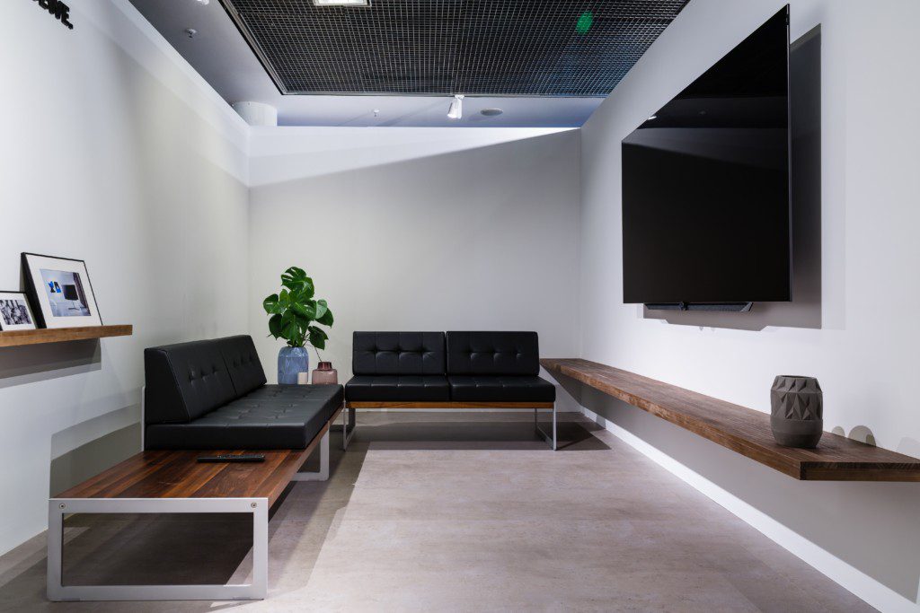 Foto de uma sala de TV moderna em tons de cinza e preto. Nela há um sofá grande em L, um hack e uma TV.