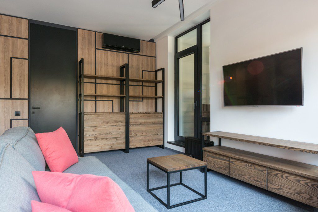 A foto mostra uma sala de TV moderna com um sofá e almofadas. Entre os móveis há uma estante grande na parede, um hack e uma mesa de centro. Todos de madeira e ferro.