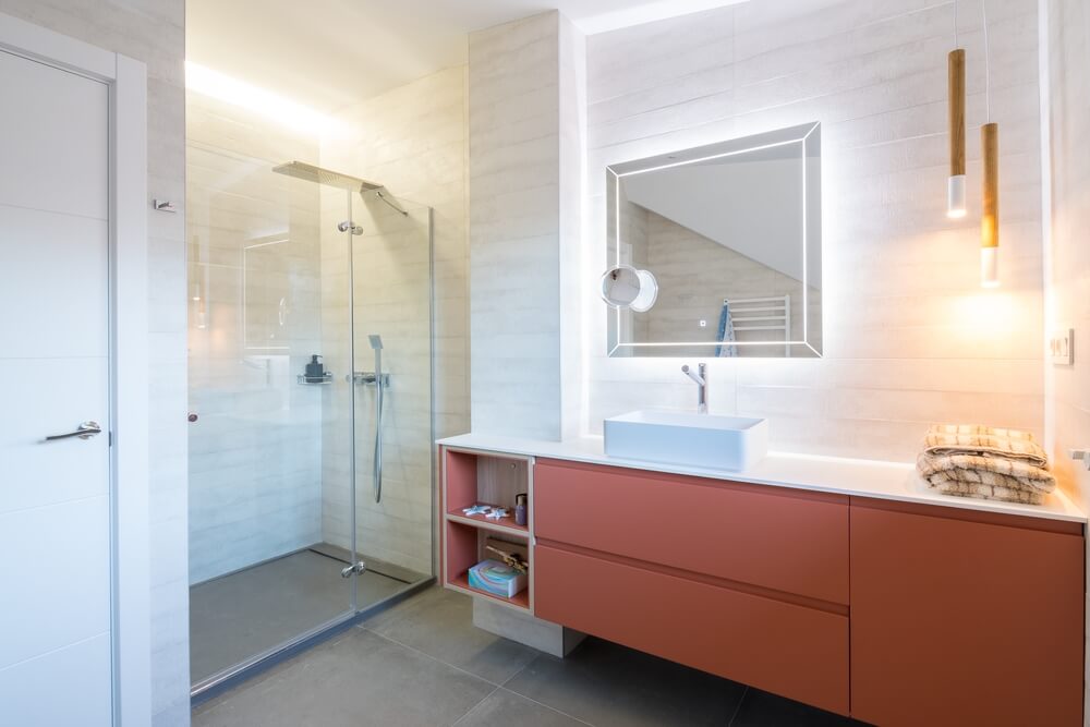 Foto que ilustra matéria sobre banheiro planejado mostra uma bancada branca de um banheiro, com uma cuba também branca para a pia. Abaixo um armário de madeira com a cor avermelhada. Acima da ía há um espelho quadrado. E do lado esquerdo um box de vidro. 