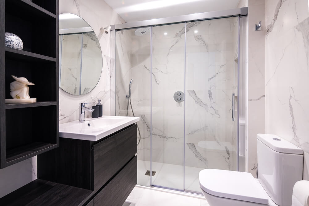 Foto que ilustra matéria sobre banheiro planejado mostra um banheiro com um box de vidro ao fundo, uma pia à esquerda, com armário de madeira abaixo, nichos ao lado e um espelho redondo acima, e à direita um vaso sanitário branco. 