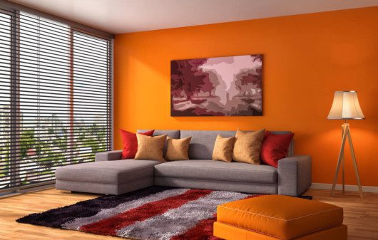 Foto que ilustra matéria sobre combinação de cores mostra uma sala de estar com uma parede laranja, um sofá cinza e almofadas vermelhas e amarelas em cima. Em primeiro plano, aparece um pufe também na cor laranja e um tapete cinza com listras vermelhas e pretas.