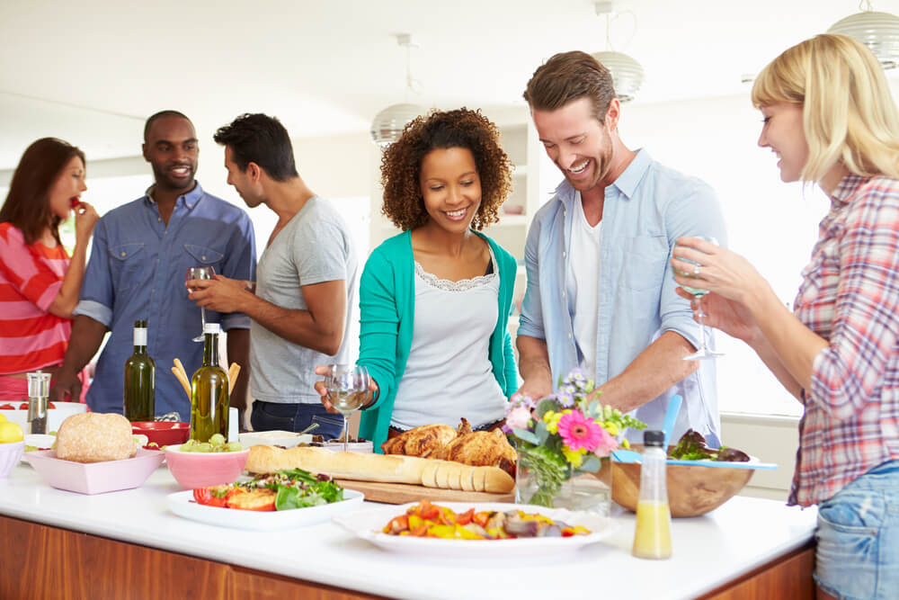 Foto que ilustra matéria sobre decoração de mesa mostra pessoas reunidas ao redor de uma mesa repleta de comidas em uma festa. 