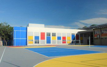 Foto que ilustra matéria sobre escolas em Canoas mostra um dos pátios do Colégio La Salle Niterói, com uma quadra poliesportiva em primeiro plano e um prédio branco e baixo ao fundo, com detalhes coloridos em vermelho, azul e amarelo, em um dia ensolarado de céu azul.