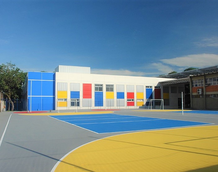 Foto que ilustra matéria sobre escolas em Canoas mostra um dos pátios do Colégio La Salle Niterói, com uma quadra poliesportiva em primeiro plano e um prédio branco e baixo ao fundo, com detalhes coloridos em vermelho, azul e amarelo, em um dia ensolarado de céu azul.