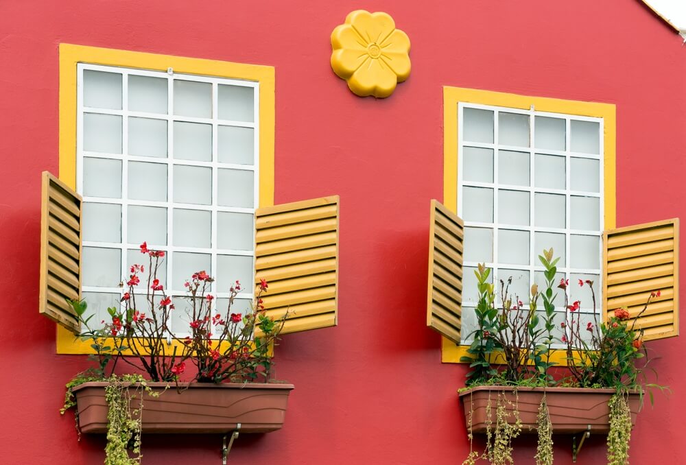 Foto que ilustra matéria sobre frente de casa simples mostra uma fachada com parede vermelha e janelas amarelas com jardineiras.