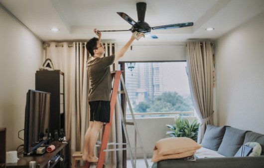Imagem de um homem em cima de uma escada limpando um ventilador de teto com um pano.