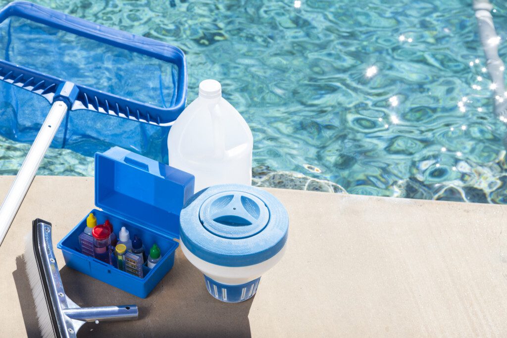 Foto que ilustra matéria sobre como limpar piscina mostra os itens necessários para fazer a limpeza da piscina, como a mangueira, a escova macia e a peneira que estão em cima da borda de uma piscina.