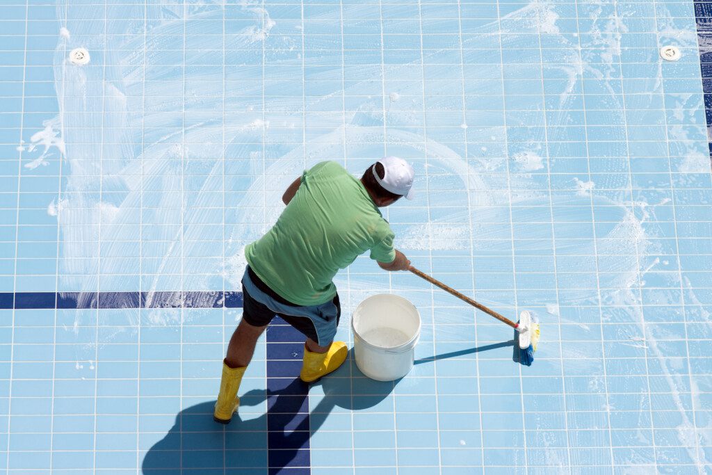Foto que ilustra matéria sobre como limpar piscina mostra um homem lavando a parte interna da piscina com uma vassoura.