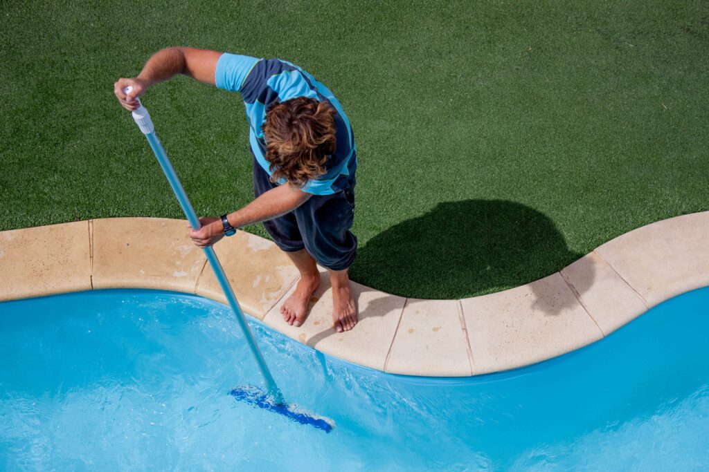 Foto que ilustra matéria sobre como limpar piscina mostra um homem segurando uma vassoura e fazendo a limpeza externa e interna de uma piscina.