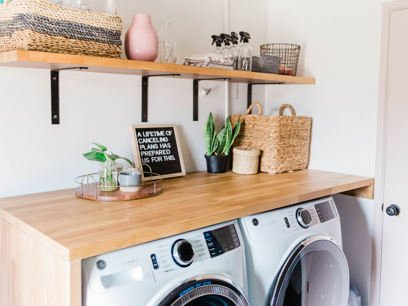 Foto de lavanderia no quintal com móveis como mesa, prateleiras e objetos de decoração. Máquina de lavar no centro. Foto disponível no Canva