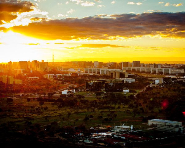 Foto que ilustra matéria sobre morar em Brasília mostra a cidade em uma panorâmica grande e vista do alto, na Esplanada dos Ministérios do centro para a direita da tela, onde se encontram as torres do Congresso Nacional. Do meio para baixo, áreas gramadas e baixas. E ao fundo, o sol baixo ilumina a paisagem.