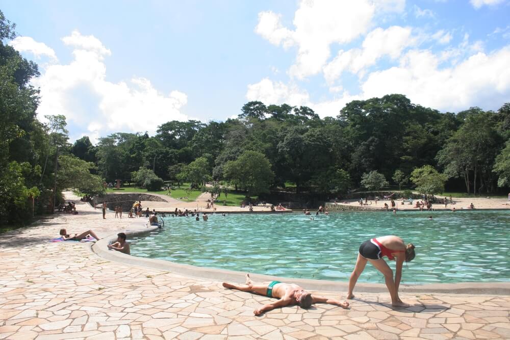 Foto que ilustra matéria sobre Parques em Brasília mostra uma das piscinas do Parque Nacional de Brasília com pessoas dentro da água ou relaxando do lado de fora. Ao fundo, muitas árvores. 