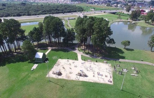 Foto que ilustra matéria sobre parques em São José dos Pinhais mostra a visão aérea do parque municipal da cidade, com um espaço de areia dentro de um gramado, onde se encontram aparelhos de ginástica, árvores e um lago mais ao fundo.