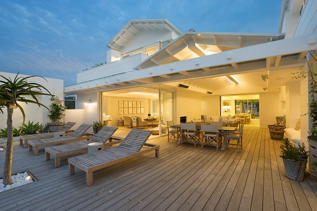 Área gourmet externa branca, com piso de madeira, cadeiras de praia e iluminação amarela. Imagem disponível em Unsplash.