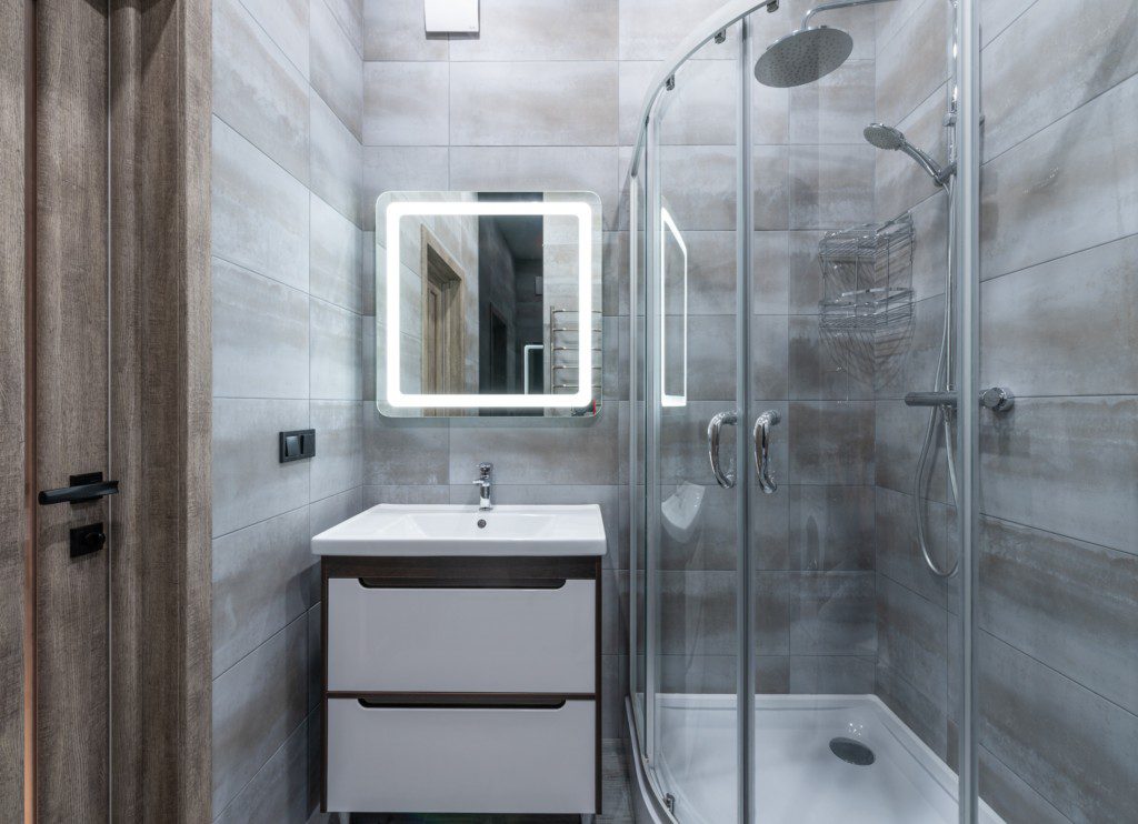 Foto de um banheiro moderno. Nele há um espelho quadrado com luz em volta, uma pia com armário embutido e um box todo transparente. Dentro do box há um chuveiro e uma ducha.