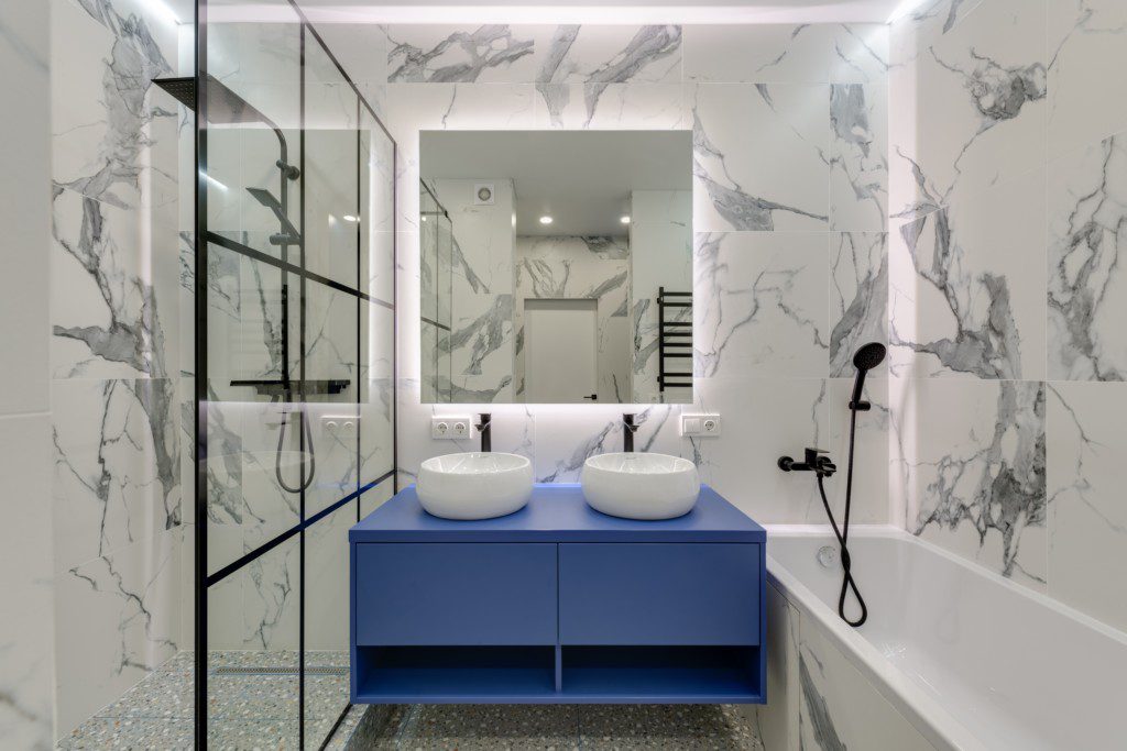 Foto de um banheiro moderno onde há um destaque para a cor azul do armário. Há também um espelho quadrado com iluminação especial, além de um box com chuveiro e ducha. O cômodo tem ainda uma banheira retangular branca.