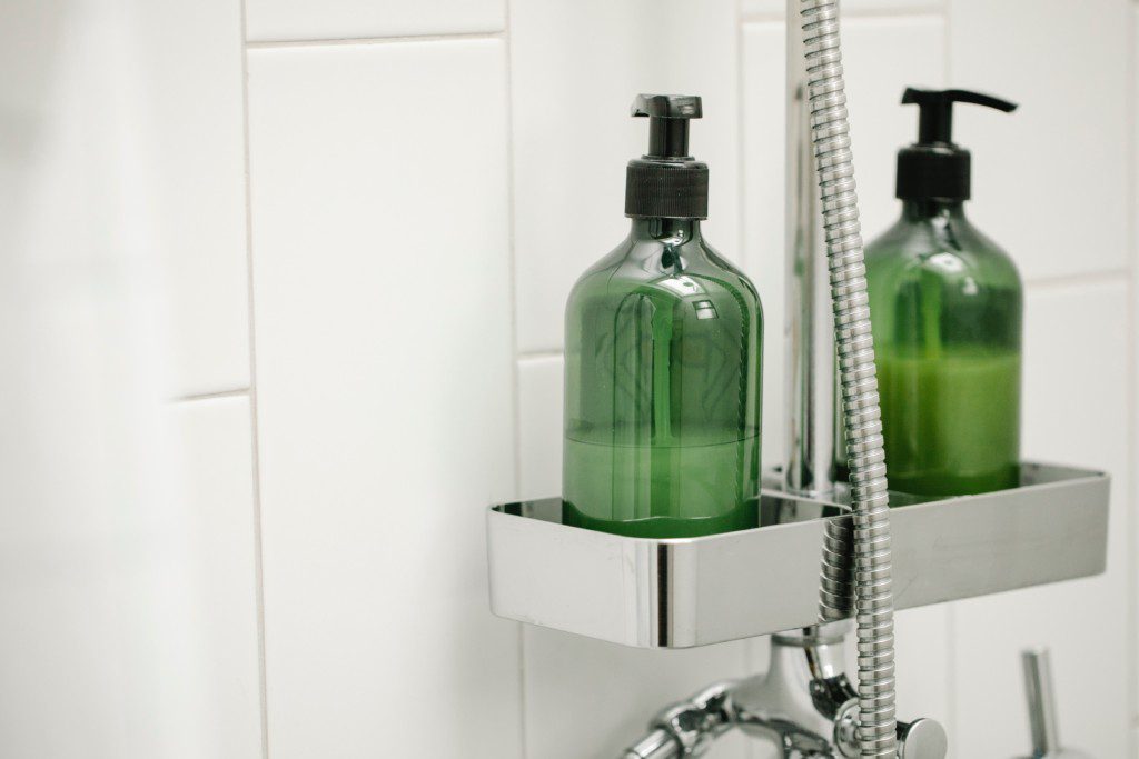Foto de um box em um banheiro moderno onde mostra um porta shampoo acoplado aos canos de inox da ducha. Nele há também dois vidros de shampoo e/ou creme para cabelo.