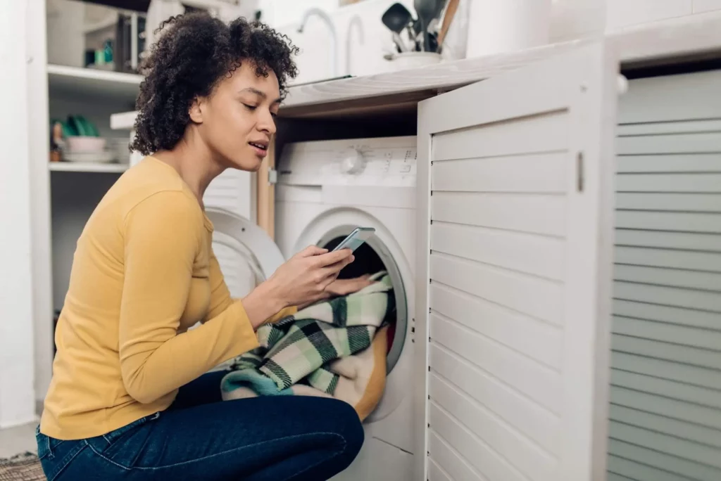 Imagem de uma mulher colocando roupas para lavar enquanto lê alguma informação no celular para ilustrar matéria sobre como limpar máquina de lavar com vinagre e bicarbonato
