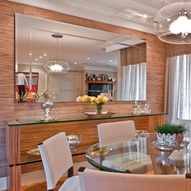 Foto de um exemplo de uma sala de jantar em um apartamento pequeno decorado. A imagem mostra uma parede com um espelho bem grande. Abaixo dele está uma estante de vidro. Há também uma mesa de vidro com cadeiras estofadas ao redor.