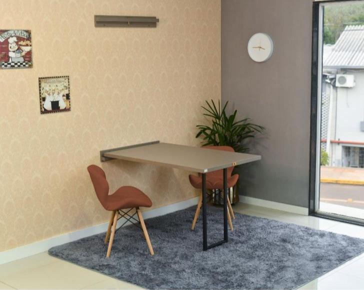 Foto de um exemplo de uma sala de jantar em um apartamento pequeno decorado. A imagem mostra uma mesa com duas cadeiras. Há também na imagem quadros decorativos, um relógio de parede e um tapete.
