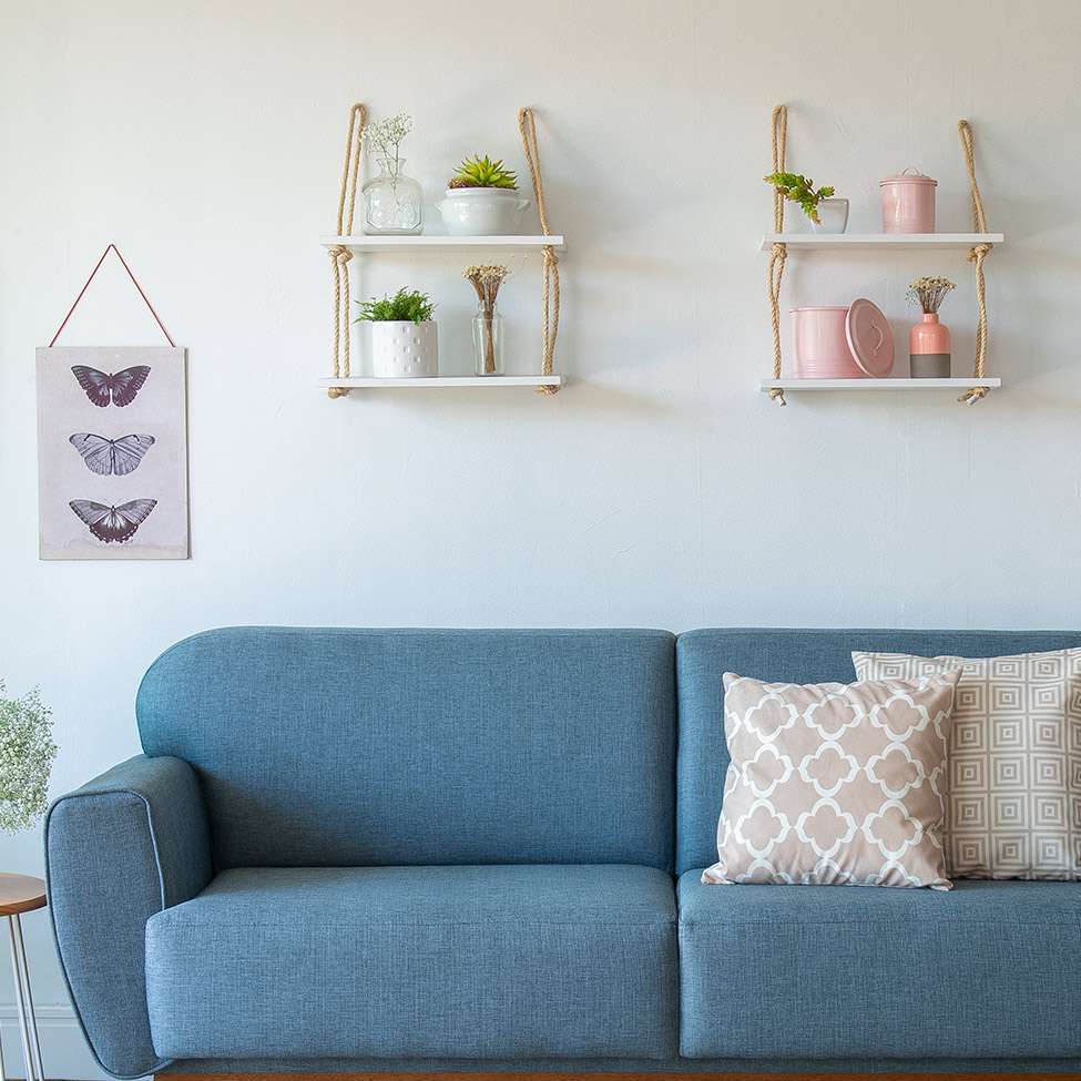 Foto de um exemplo de uma sala de estar em um apartamento pequeno decorado. A imagem mostra um sofá azul com duas almofadas. Acima dele há duas prateleiras pendentes com itens como plantas e potes decorativos. Há também um quadro decorativo ao lado do sofá.
