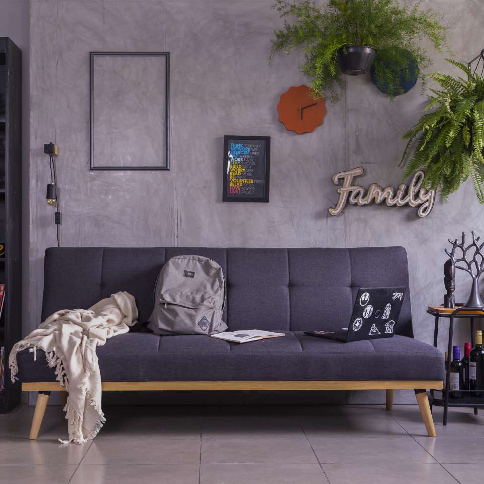 Foto de um exemplo de uma sala de estar em um apartamento pequeno decorado. A imagem mostra um sofá azul com mochila, manta e notebook em cima dele. Há também quadros decorativos e plantas ao redor.