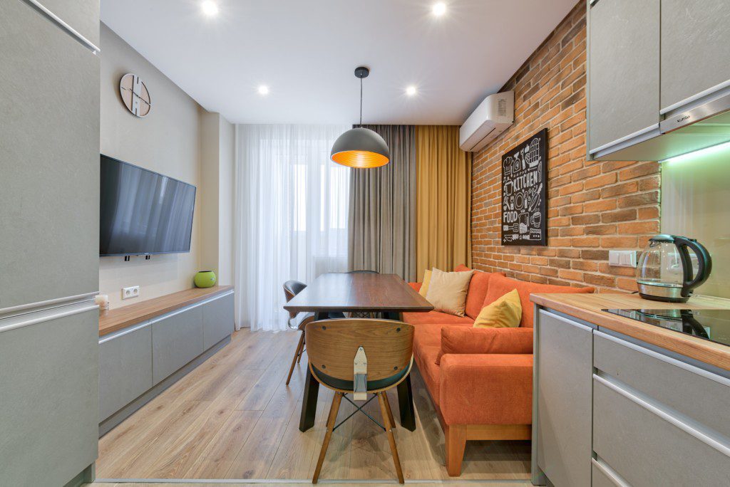 A foto mostra um exemplo de decoração retrô em um studio com bancada de cozinha, sofá laranja, mesa retangular com duas cadeiras, rack e TV.