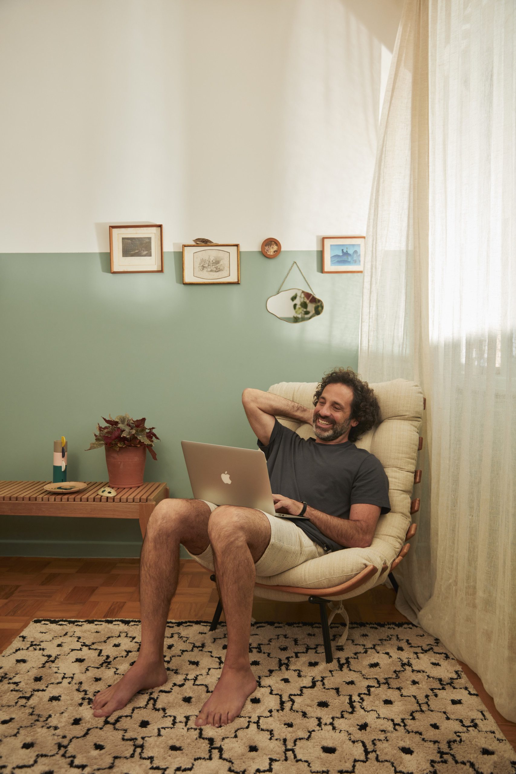 A foto mostra um exemplo de decoração retrô que usa quadros de memórias afetivas na parede. Há também: uma mesa lateral, um homem sentado na poltrona com um notebook, tapete e cortina.
