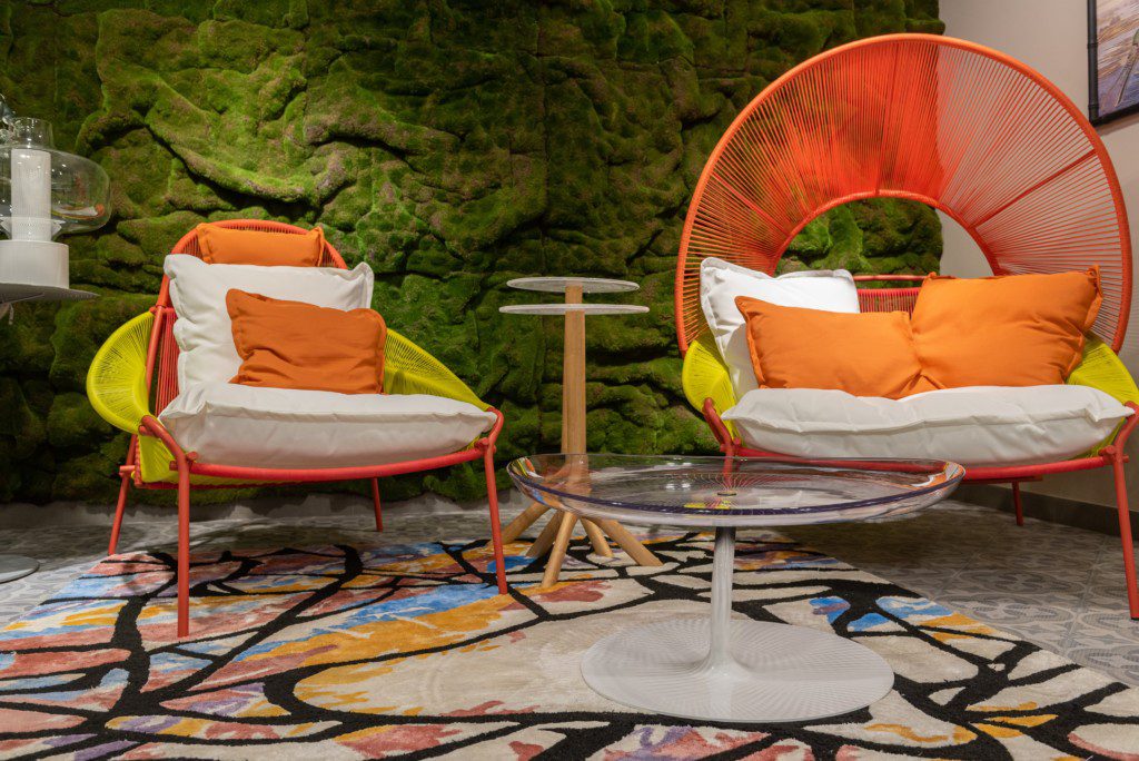  A foto mostra um exemplo de decoração retrô em uma sala de estar com duas poltronas laranjas, uma mesa de centro, um tapete colorido e uma parede verde.