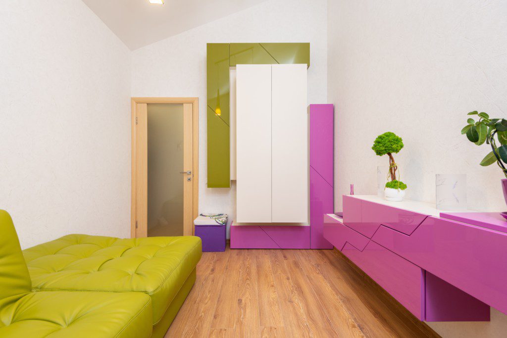 A foto mostra um exemplo de decoração retrô na sala de estar com sofá verde, estante rosa, armário branco e plantas decorativas.