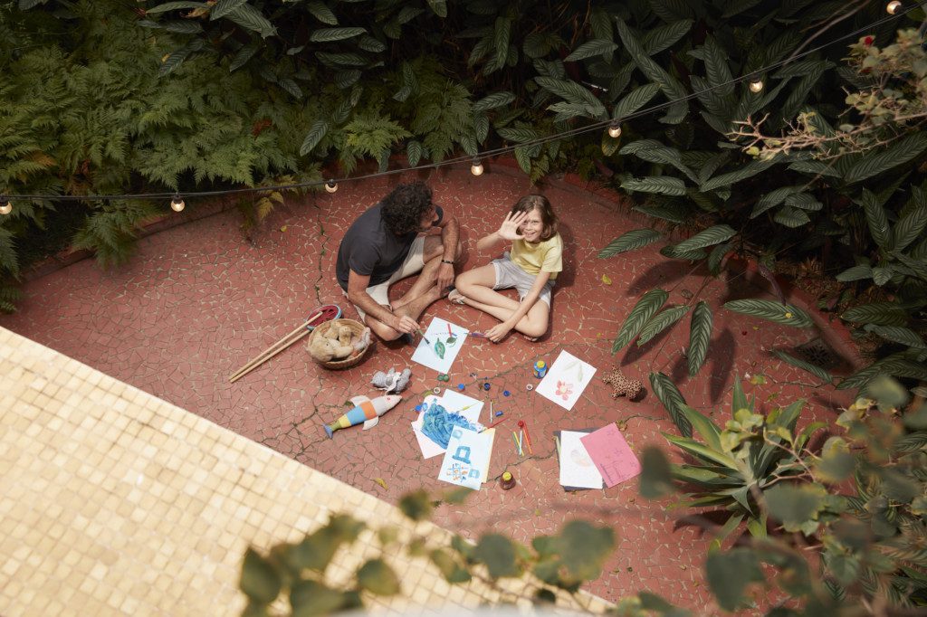 A foto mostra um exemplo de decoração retrô com varal de luzes em um quintal. No chão, há uma criança e um adulto pintando. Ao redor deles, muitas plantas.
